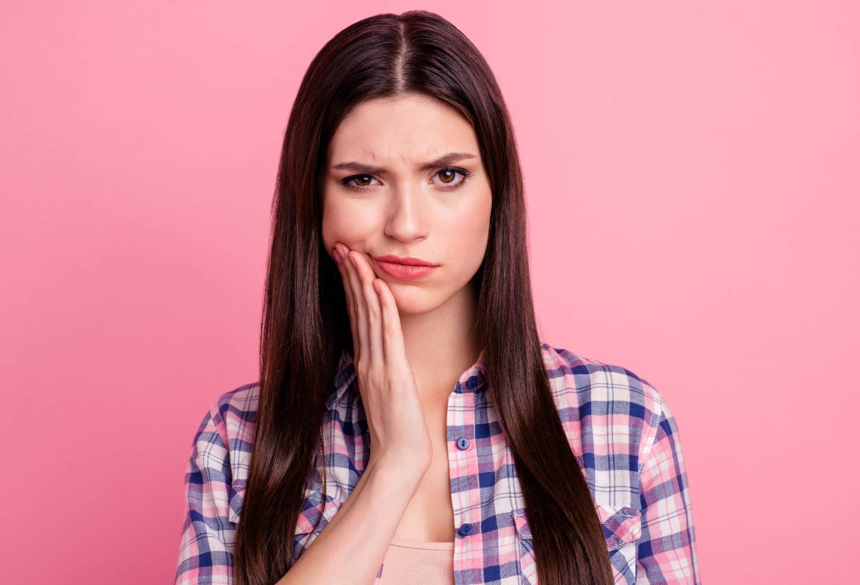 Brunette woman with sensitive teeth cringes in pain and touches her cheek against a pink background