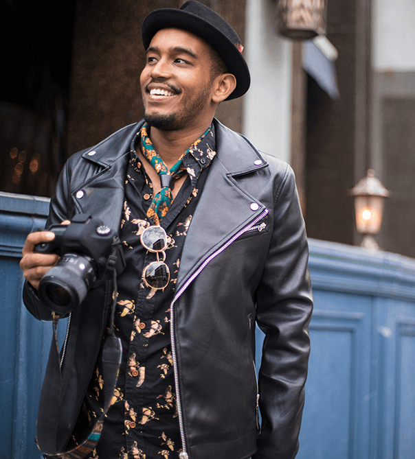 black man in leather jacket and fedora smiling and holding a camera