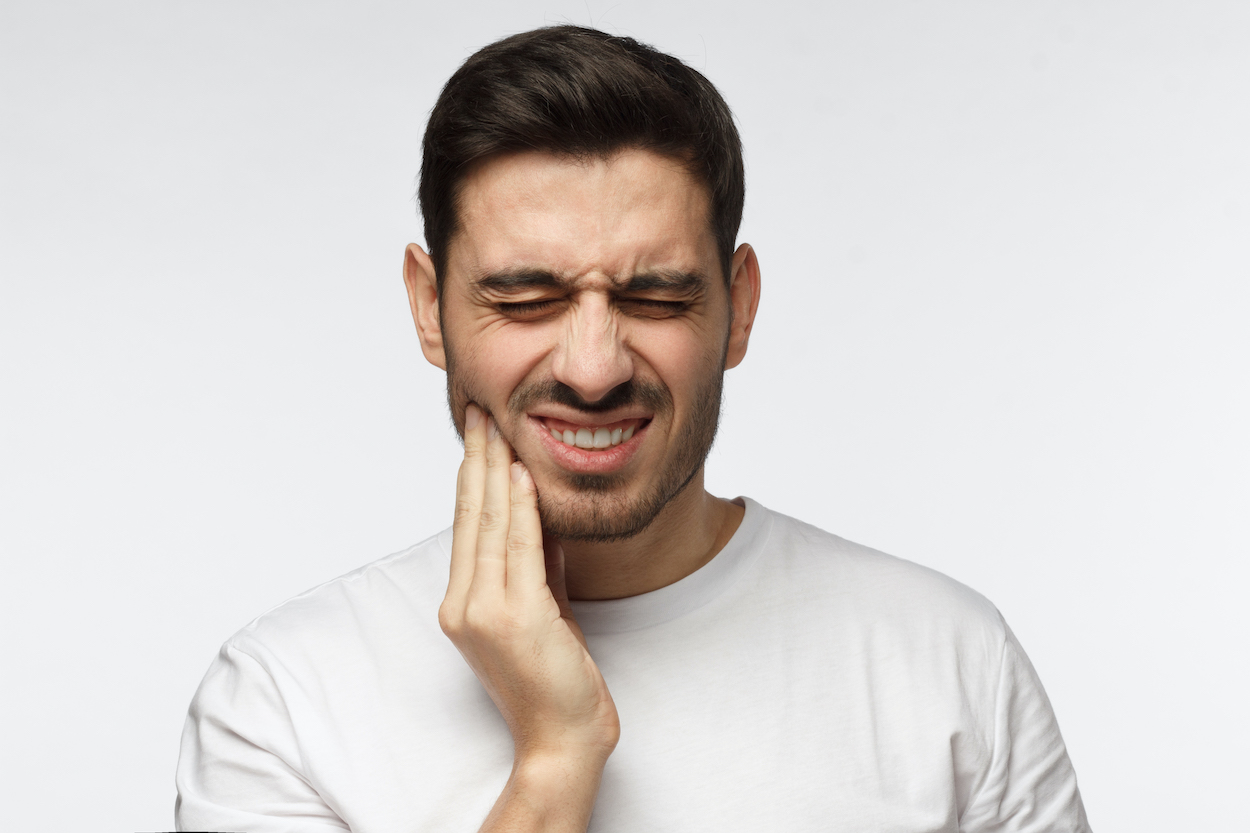 Brunette man in a white shirt touches his cheek and cringes in pain due to an untreated cavity