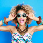 Happy Black woman with sunglasses and summer clothes smiles against a blue wall