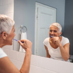 Older woman with white hair looks in the bathroom mirror as she brushes her teeth